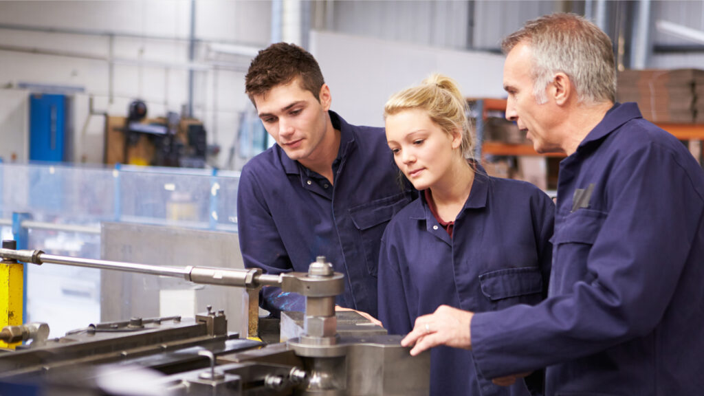 Zwei Jugendliche und ein Erwachsener in Arbeitskleidung stehen in einer Werkstatt. Der Mann erklärt den Jugendlichen eine Produktionsmaschine.