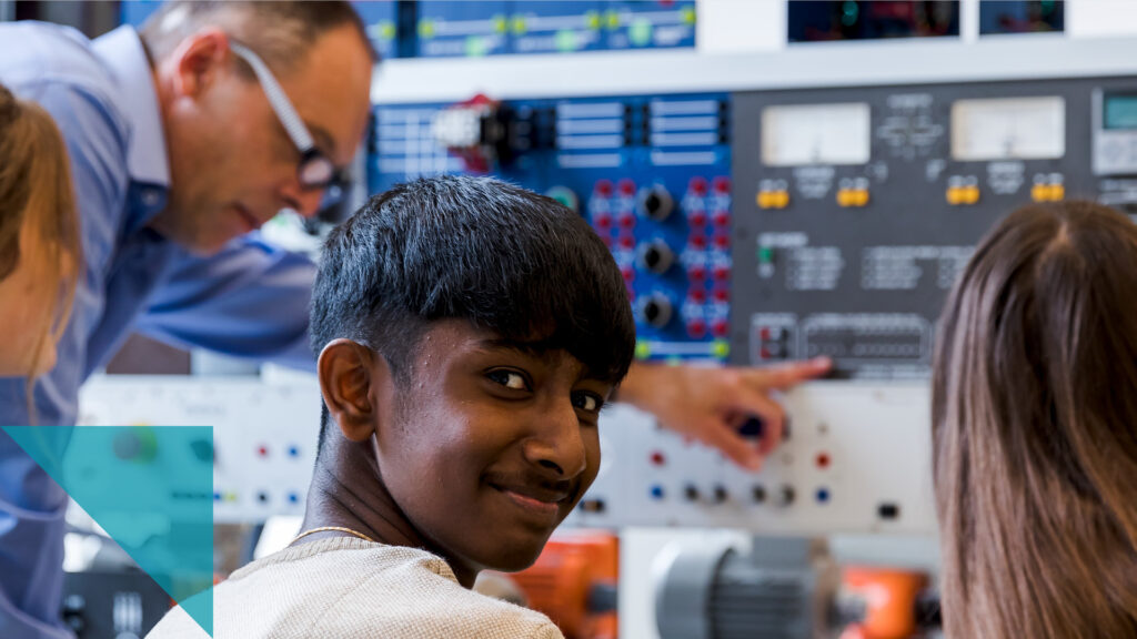 In einer Werkstatt erklärt ein Erwachsener den Lernenden ein elektronisches Board, ein Schüler schaut über die Schulter zurück in die Kamera und lächelt.