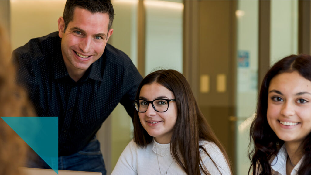 Ein Lehrer und zwei jugendliche Schülerinnen in einem Schulzimmer lächeln in die Kamera.