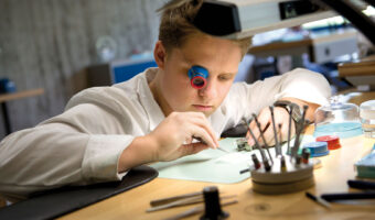 Ein Mann mit einer Monokellupe am Auge arbeitet an einem Uhrwerk.