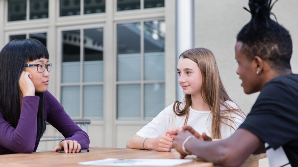 Zwei Schülerinnen und ein Schüler tauschen sich am Tisch sitzend aus