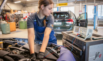 Eine Frau prüft, im Motorraum eines Personenwagens, eine Funktion mit einem Messgerät.