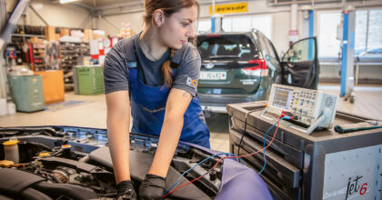 Eine Frau prüft, im Motorraum eines Personenwagens, eine Funktion mit einem Messgerät.